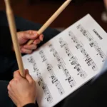 A close-up of a drummer's hands holding drum sticks, poised above a sheet of drum cadence music